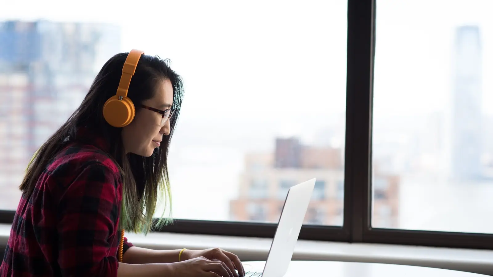 A woman wearing headphones at a laptop beside a window