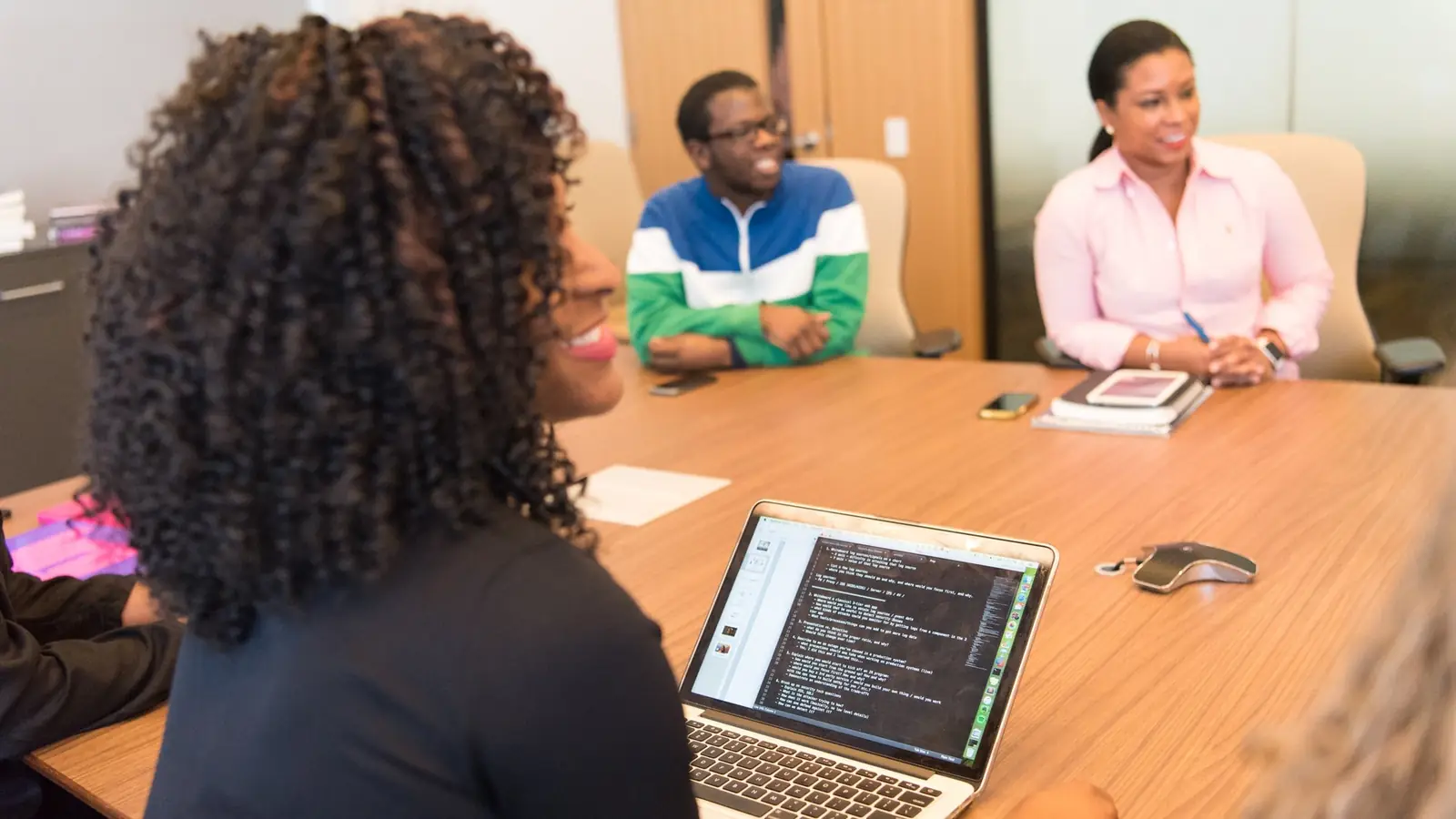 A woman works at a laptop while colleagues sit around a conference table